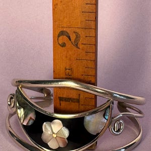 May include: A silver-toned cuff bracelet with a black inlay, featuring a flower and abalone shell accents. The bracelet is approximately 1 inch wide. A wooden ruler is placed behind the bracelet for scale.