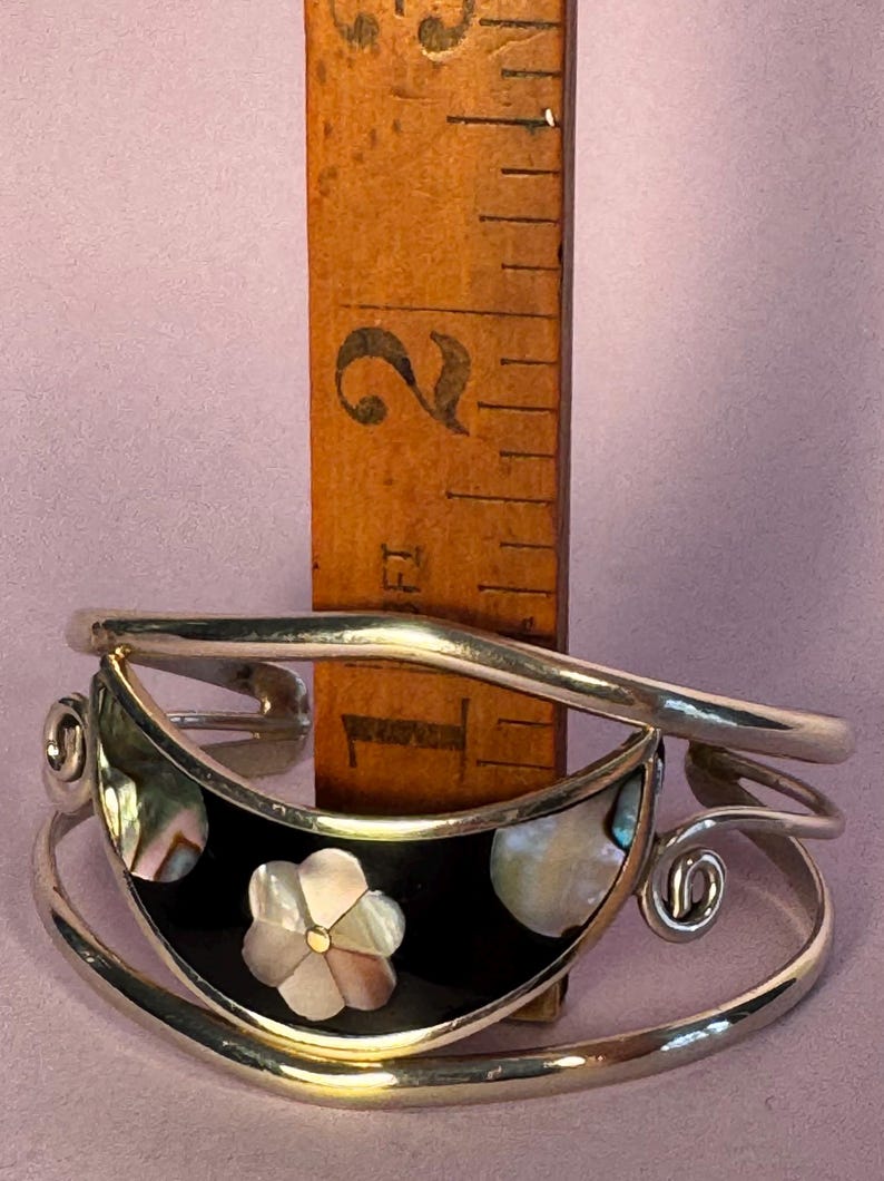 May include: A silver-toned cuff bracelet with a black inlay, featuring a flower and abalone shell accents. The bracelet is approximately 1 inch wide. A wooden ruler is placed behind the bracelet for scale.
