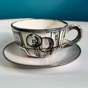 Puede incluir: Un juego de taza y platillo de cerámica. La taza y el platillo son blancos con un borde negro. La taza tiene un diseño abstracto negro. El asa es negra. El platillo es redondo y tiene un borde negro. El juego está sobre una superficie blanca.