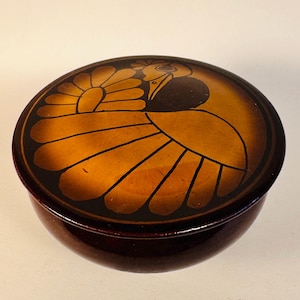 May include: A circular, dark brown decorative box with a lid. The lid displays a stylized bird design in gold and black, with a detailed feather pattern. The box has a glossy finish.