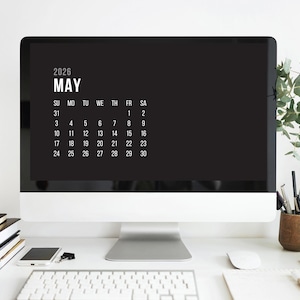May include: A computer screen shows a May 2026 calendar in white text against a black background. The computer sits on a white desk with a keyboard, mouse, and stationery. A small plant in a glass bottle and other items complete the workspace.
