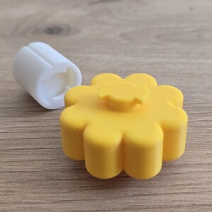 K&ouml;nnte beinhalten: Ein gelber, blumenf&ouml;rmiger Knopf mit einem zentralen Vorsprung und ein separates wei&szlig;es zylindrisches Bauteil mit einer geschlitzten &Ouml;ffnung. Das gelbe Teil hat ein gewelltes Design, w&auml;hrend das wei&szlig;e Teil glatt und an einem Ende hohl ist.