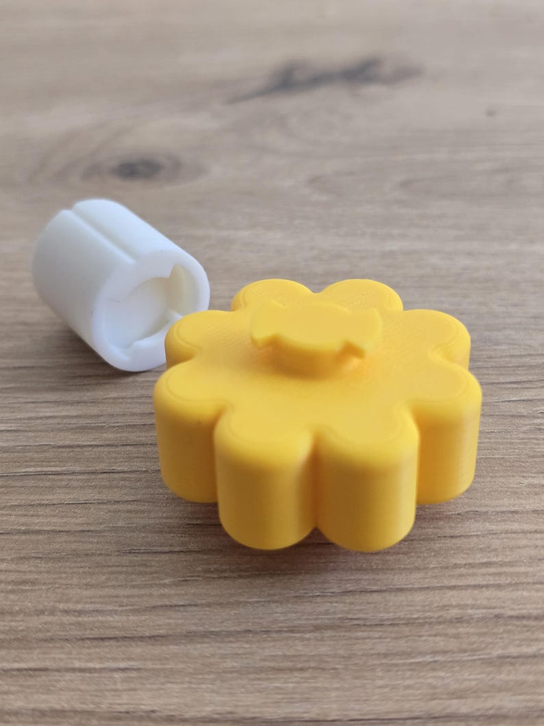 K&ouml;nnte beinhalten: Ein gelber, blumenf&ouml;rmiger Knopf mit einem zentralen Vorsprung und ein separates wei&szlig;es zylindrisches Bauteil mit einer geschlitzten &Ouml;ffnung. Das gelbe Teil hat ein gewelltes Design, w&auml;hrend das wei&szlig;e Teil glatt und an einem Ende hohl ist.