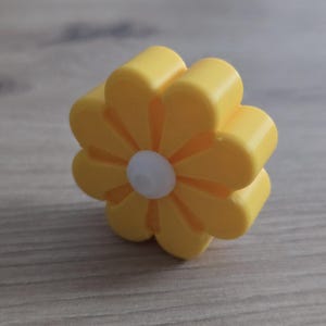 K&ouml;nnte beinhalten: Ein kleines, dreidimensionales blumenf&ouml;rmiges Objekt mit acht abgerundeten gelben Bl&uuml;tenbl&auml;ttern und einer kreisf&ouml;rmigen wei&szlig;en Mitte. Der Gegenstand hat eine glatte, matte Oberfl&auml;che und ein dickes, blockartiges Profil, das aufrecht steht.