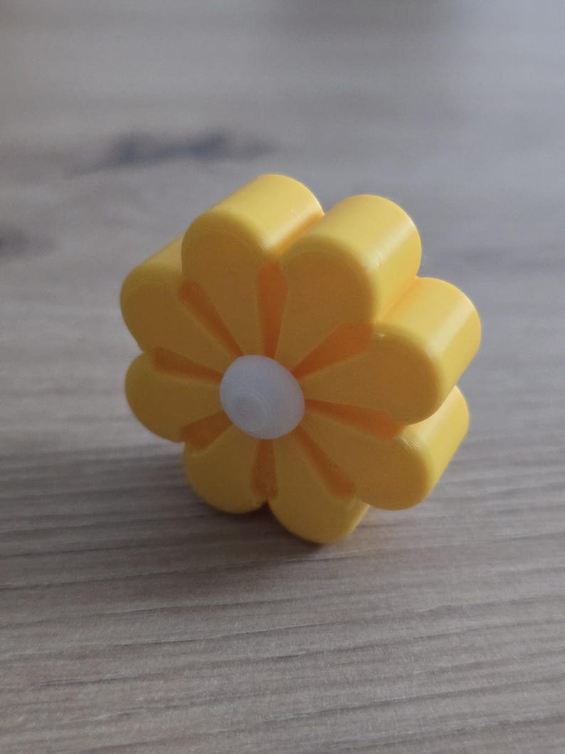 K&ouml;nnte beinhalten: Ein kleines, dreidimensionales blumenf&ouml;rmiges Objekt mit acht abgerundeten gelben Bl&uuml;tenbl&auml;ttern und einer kreisf&ouml;rmigen wei&szlig;en Mitte. Der Gegenstand hat eine glatte, matte Oberfl&auml;che und ein dickes, blockartiges Profil, das aufrecht steht.