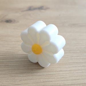K&ouml;nnte beinhalten: Ein dreidimensionales, blumenf&ouml;rmiges Objekt mit acht abgerundeten wei&szlig;en Bl&uuml;tenbl&auml;ttern und einer kleinen, kreisf&ouml;rmigen, erhabenen gelben Mitte. Der Gegenstand hat eine glatte, matte Oberfl&auml;che und ein dickes, gleichm&auml;&szlig;iges Profil.