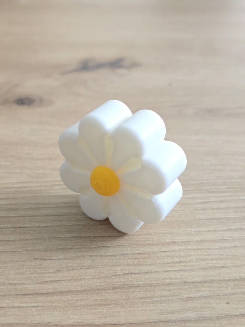 K&ouml;nnte beinhalten: Ein dreidimensionales, blumenf&ouml;rmiges Objekt mit acht abgerundeten wei&szlig;en Bl&uuml;tenbl&auml;ttern und einer kleinen, kreisf&ouml;rmigen, erhabenen gelben Mitte. Der Gegenstand hat eine glatte, matte Oberfl&auml;che und ein dickes, gleichm&auml;&szlig;iges Profil.