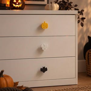 May include: A white three-drawer dresser featuring three distinct decorative knobs. The top knob is a yellow pumpkin shape, the middle knob is a white ghost shape, and the bottom knob is a black bat shape, all attached to the drawer fronts.