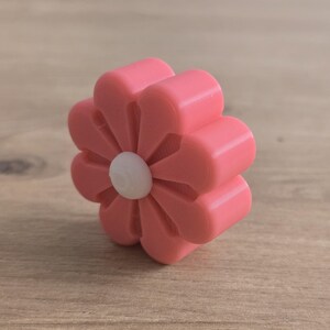 K&ouml;nnte beinhalten: Ein rosafarbener, blumenf&ouml;rmiger Knopf mit acht abgerundeten Bl&uuml;tenbl&auml;ttern und einem kleinen, kreisf&ouml;rmigen wei&szlig;en Mittelst&uuml;ck. Das Objekt hat eine glatte, matte Oberfl&auml;che und eine dreidimensionale Struktur mit sichtbaren Schichttexturen.