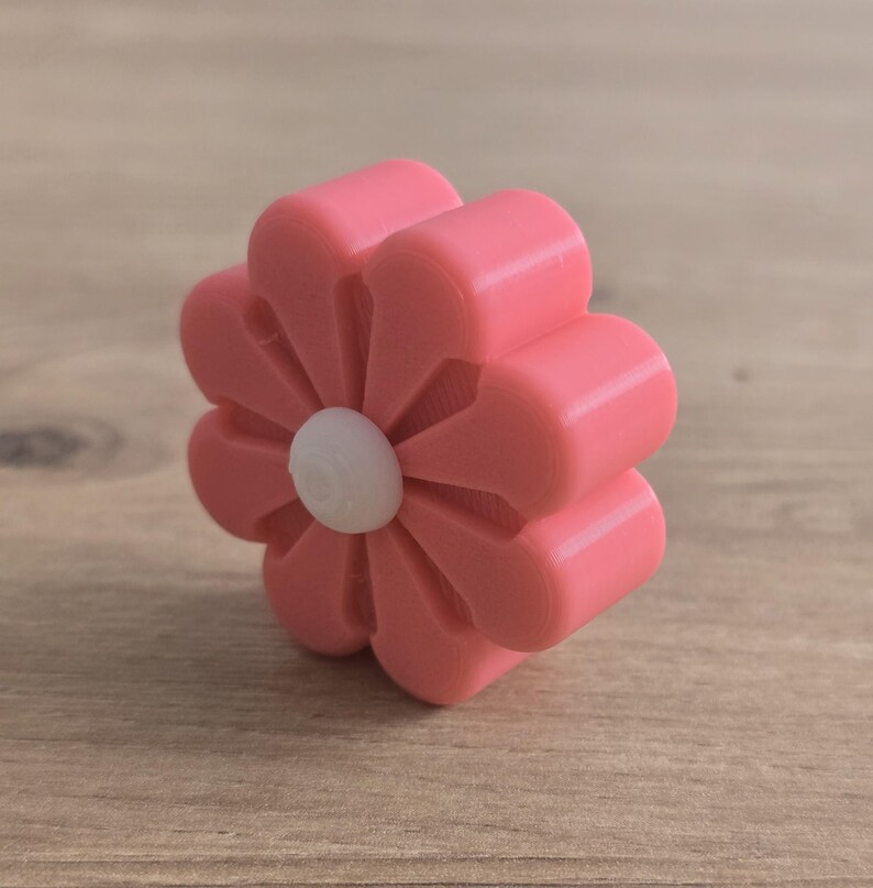 K&ouml;nnte beinhalten: Ein rosafarbener, blumenf&ouml;rmiger Knopf mit acht abgerundeten Bl&uuml;tenbl&auml;ttern und einem kleinen, kreisf&ouml;rmigen wei&szlig;en Mittelst&uuml;ck. Das Objekt hat eine glatte, matte Oberfl&auml;che und eine dreidimensionale Struktur mit sichtbaren Schichttexturen.