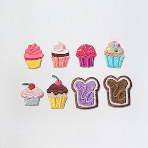 May include: A collection of embroidered patches depicting cupcakes and toast. The cupcakes feature pink, brown, and blue frosting with sprinkles and cherries. The toast slices are purple and brown with a textured design. Ideal for customising clothing or bags.