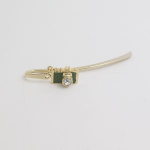 May include: A gold bookmark with a miniature camera charm. The camera has a green body, gold detailing, and a clear crystal lens. The bookmark has a curved, slender design.
