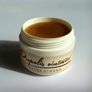 May include: A white jar of propolis ointment with a light brown cream inside. The jar has a label with the words "Propolis ointment EXTRA STRONG" in brown script. The jar is sitting on a white surface.
