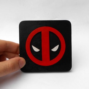 May include: Black square coaster with a red and white Deadpool logo. The logo is a circle with a red outline and a white mask inside.