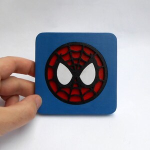 以下が含まれることがあります： 青い正方形のコースターに、赤と黒のスパイダーマンのマスクのデザインが施されています。マスクは白い目を持っています。