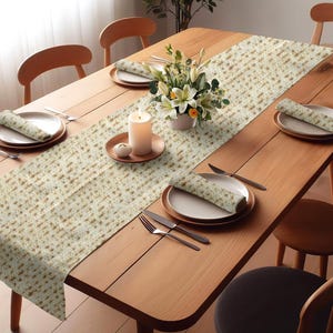 May include: A beige table runner with a matzah pattern. The runner is laid on a wooden table with place settings and a vase of white flowers.