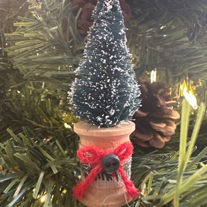 May include: A miniature Christmas tree ornament. The green tree is atop a wooden spool decorated with a red bow and a gray button. The ornament is set against a backdrop of a Christmas tree with pine needles and a pine cone.