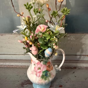 May include: A decorative floral arrangement in a white pitcher with a light blue base. The pitcher is adorned with pink and orange floral designs. The arrangement includes a variety of artificial flowers, buds, and greenery, along with a small pink pig and a striped Easter egg.