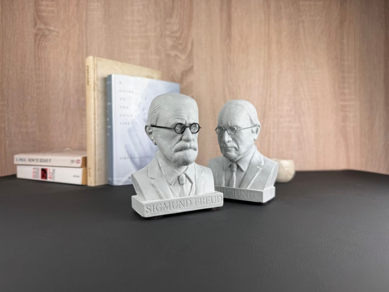 May include: Two grey busts of Sigmund Freud and Carl Jung, each wearing spectacles. The Freud bust is labelled with his name. Books and a small decorative object are in the background.