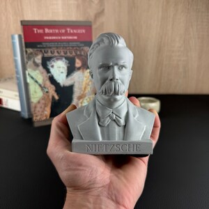 May include: A gray bust of Friedrich Nietzsche, with the name "NIETZSCHE" inscribed on the base. The sculpture features detailed facial features, including a prominent mustache. A copy of "The Birth of Tragedy" is visible in the background.