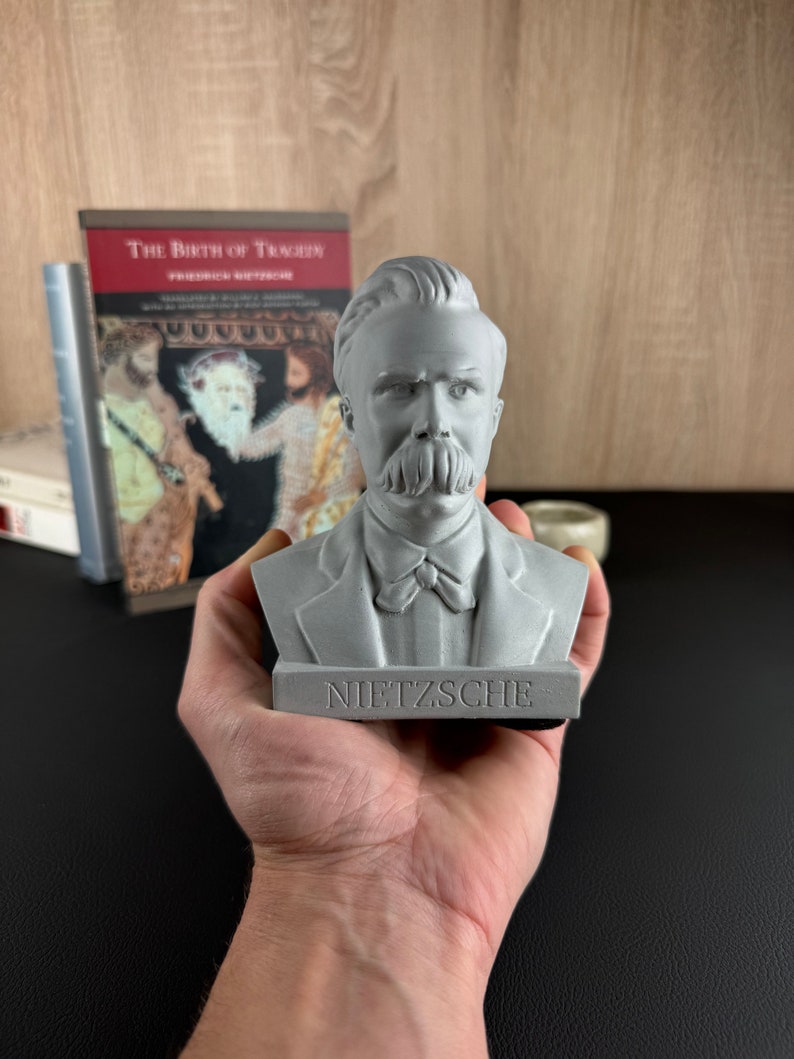 May include: A gray bust of Friedrich Nietzsche, with the name "NIETZSCHE" inscribed on the base. The sculpture features detailed facial features, including a prominent mustache. A copy of "The Birth of Tragedy" is visible in the background.