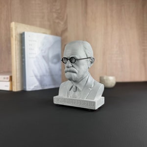 May include: A grey bust of Sigmund Freud, wearing round spectacles and a suit. The bust is on a black base with the name "SIGMUND FREUD" inscribed. Books and a small decorative item are in the background.