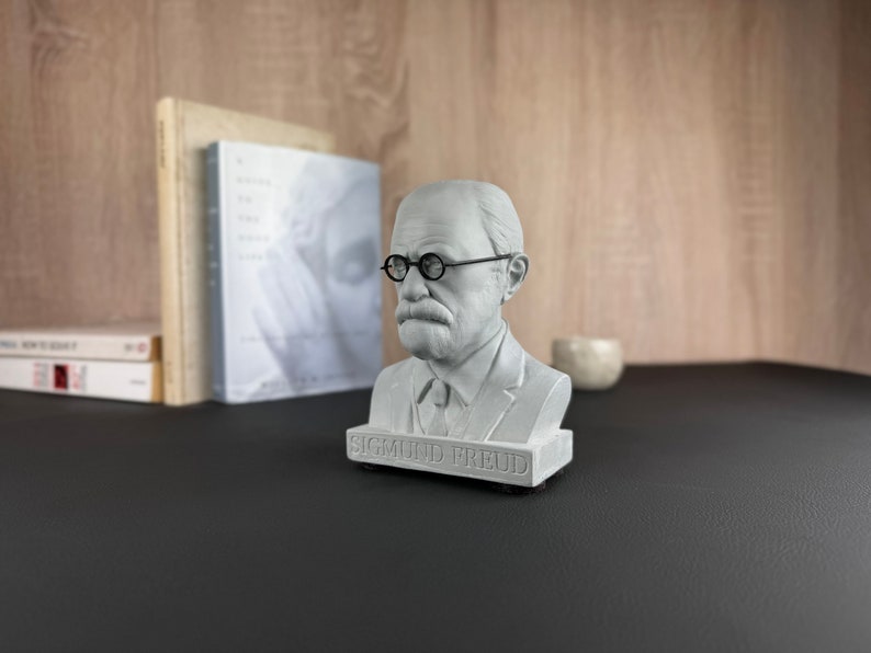 May include: A grey bust of Sigmund Freud, wearing round spectacles and a suit. The bust is on a black base with the name "SIGMUND FREUD" inscribed. Books and a small decorative item are in the background.