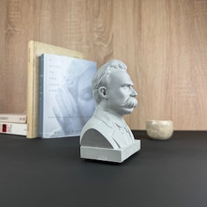 May include: A gray bust of a man with a large mustache, likely a philosopher, sits on a black surface. The bust is next to books and a small, light-colored ceramic bowl. The background is a light wood grain.
