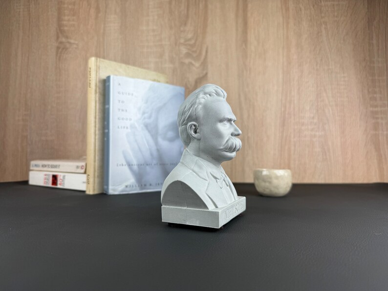 May include: A gray bust of a man with a large mustache, likely a philosopher, sits on a black surface. The bust is next to books and a small, light-colored ceramic bowl. The background is a light wood grain.