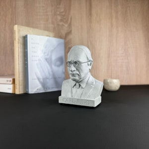 May include: A grey bust of Carl Jung, wearing spectacles and a tie, is on a black surface. Behind it are books, including one titled "A Guide to the Good Life." A small, round, light-coloured object is to the right.