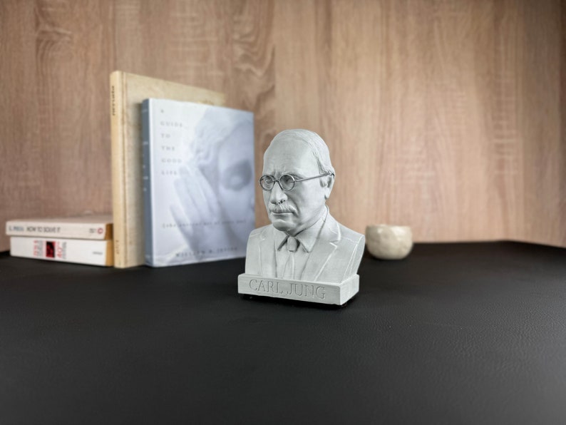 May include: A grey bust of Carl Jung, wearing spectacles and a tie, is on a black surface. Behind it are books, including one titled "A Guide to the Good Life." A small, round, light-coloured object is to the right.