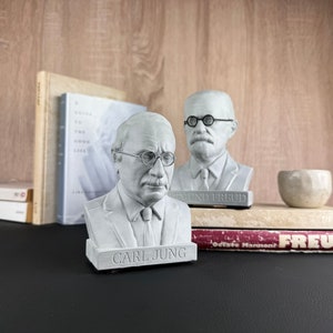 May include: Two white busts of Sigmund Freud and Carl Jung, each wearing spectacles, are displayed. The Carl Jung bust has the name "CARL JUNG" on the base. Books and a small white bowl are also on the table.