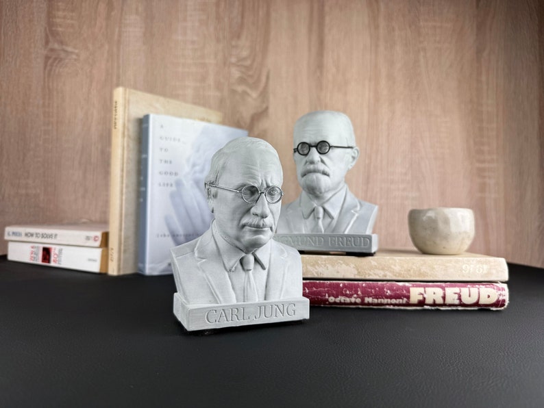 May include: Two white busts of Sigmund Freud and Carl Jung, each wearing spectacles, are displayed. The Carl Jung bust has the name "CARL JUNG" on the base. Books and a small white bowl are also on the table.