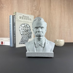 May include: A gray bust of Friedrich Nietzsche with a prominent mustache and the name "NIETZSCHE" on the base. The bust is in front of books and a small decorative bowl.