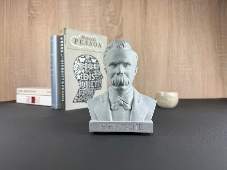 May include: A gray bust of Friedrich Nietzsche with a prominent mustache and the name "NIETZSCHE" on the base. The bust is in front of books and a small decorative bowl.