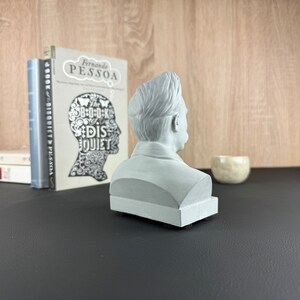 May include: A light gray bust of a man, viewed from the side, sits on a black surface. The bust is placed in front of a stack of books and a small, off-white ceramic bowl. The background features a wooden wall.