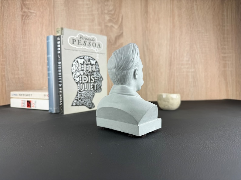 May include: A light gray bust of a man, viewed from the side, sits on a black surface. The bust is placed in front of a stack of books and a small, off-white ceramic bowl. The background features a wooden wall.