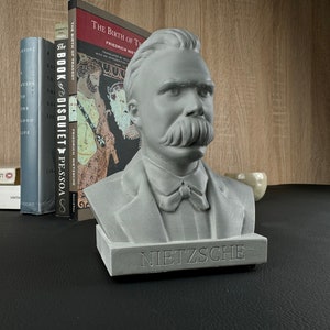 May include: A gray concrete bust of Friedrich Nietzsche with a prominent mustache and the name "NIETZSCHE" inscribed on the base. The bust is displayed on a black surface with books in the background, including "The Birth of Tragedy".