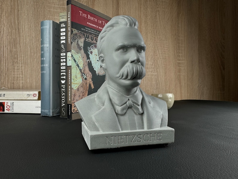 May include: A gray concrete bust of Friedrich Nietzsche with a prominent mustache and the name "NIETZSCHE" inscribed on the base. The bust is displayed on a black surface with books in the background, including "The Birth of Tragedy".