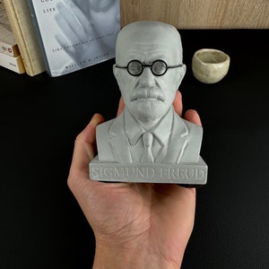 May include: A grey bust of Sigmund Freud, with round spectacles and a tie. The bust is held in a hand, with the name "SIGMUND FREUD" at the base. A book titled "Guide to the Good Life" is visible in the background.