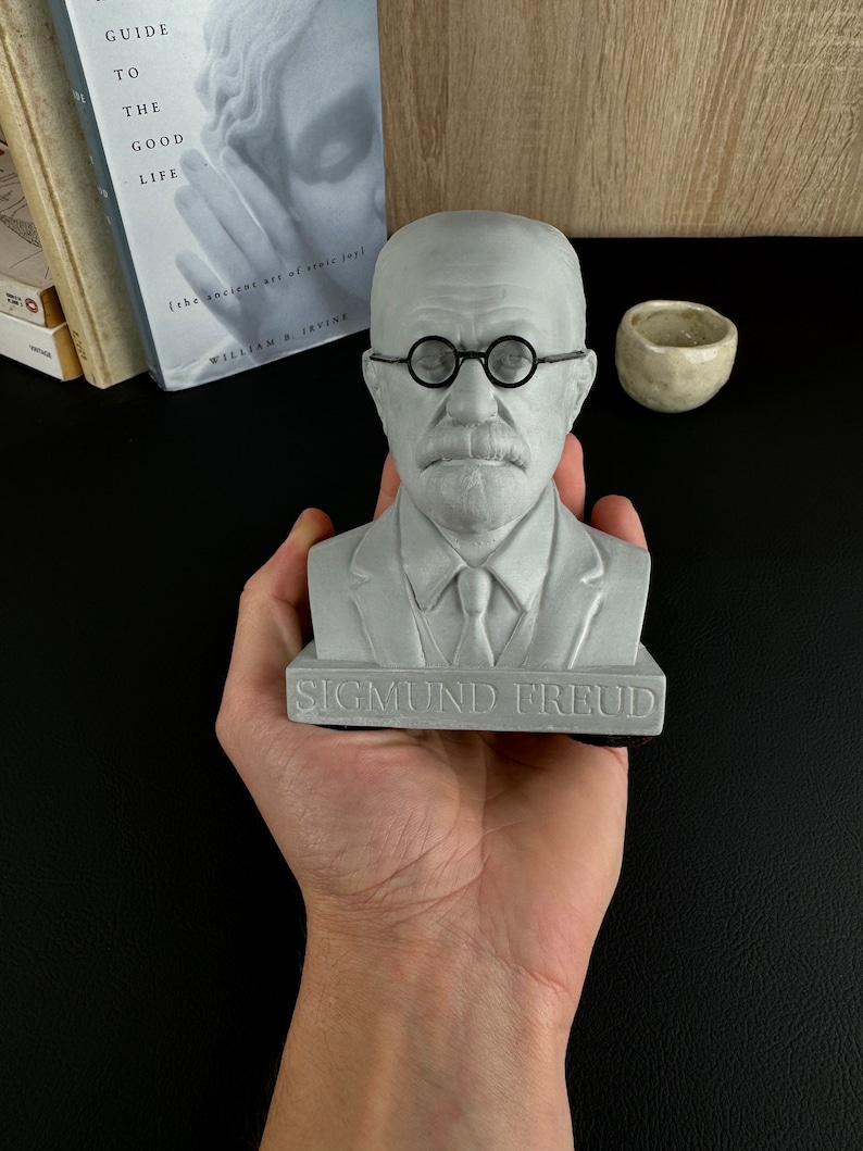 May include: A grey bust of Sigmund Freud, with round spectacles and a tie. The bust is held in a hand, with the name "SIGMUND FREUD" at the base. A book titled "Guide to the Good Life" is visible in the background.