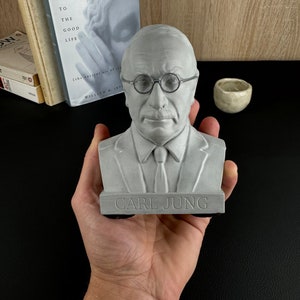 May include: A grey bust of Carl Jung, wearing round spectacles and a tie, is held in a hand. The base of the bust is inscribed with the name "CARL JUNG". Books and a small ceramic bowl are in the background.