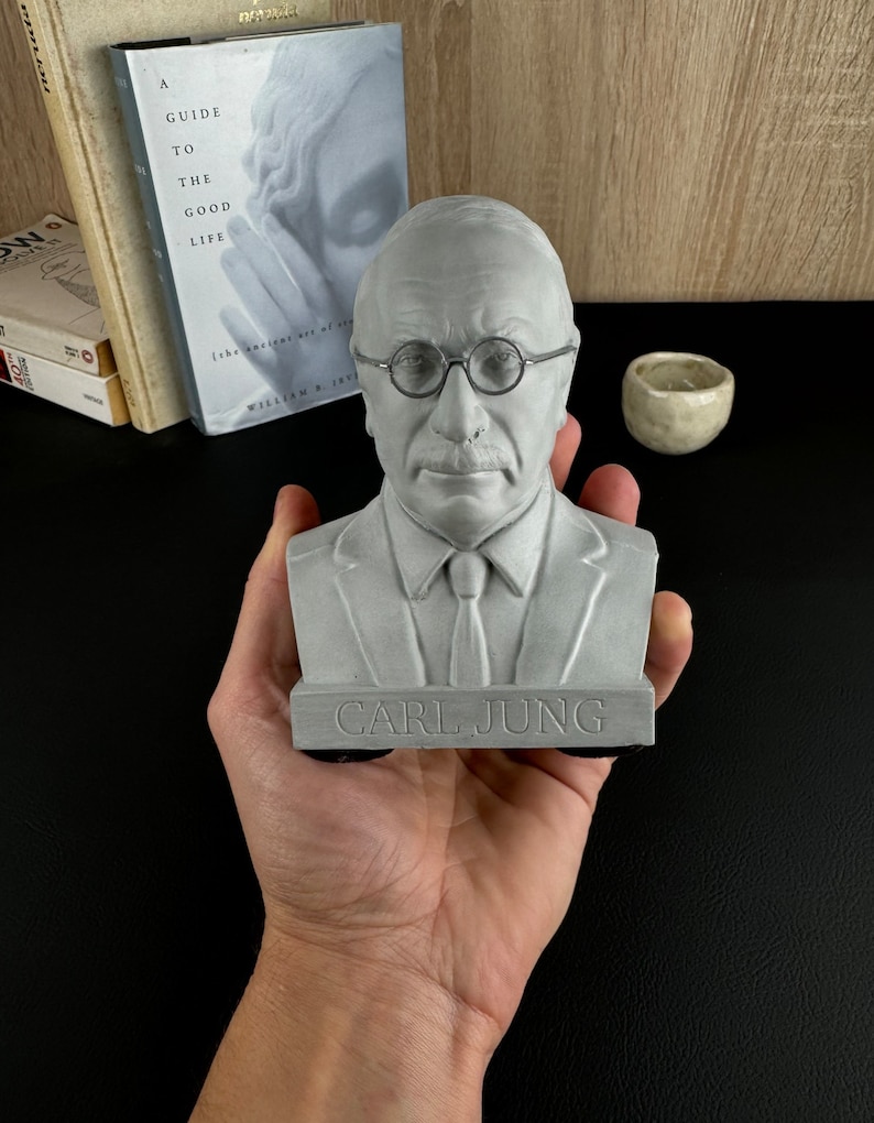 May include: A grey bust of Carl Jung, wearing round spectacles and a tie, is held in a hand. The base of the bust is inscribed with the name "CARL JUNG". Books and a small ceramic bowl are in the background.