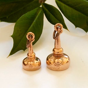 May include: Two gold-colored bottle stopper charms. The larger charm is about 1.5 inches tall and the smaller charm is about 1 inch tall. Both charms have a round base and a long, thin neck.