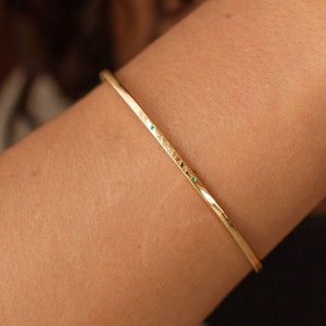 A gold bangle bracelet with a small emerald green gemstones embedded along its length. The minimalist design is simple and elegant, suitable for everyday wear. The bracelet is shown on a wrist, highlighting its delicate and comfortable fit.