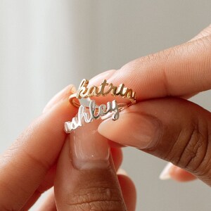 May include: Two stacked rings made of metal, one gold-toned and one silver-toned. Each ring features a name written in a cursive script font. The top ring displays the name Katrin in gold, and the bottom ring displays the name Ashley in silver.