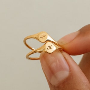May include: Two gold-toned signet rings with smooth, polished bands. Each ring features a small, oval-shaped face engraved with a single cursive letter. One ring displays the letter L, while the other ring displays the letter K.