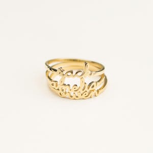 Puede incluir: Un anillo de metal dorado compuesto por tres bandas apiladas, cada una con un nombre diferente en letra cursiva. Los nombres visibles en las bandas son Zach, Amelia y Owen, integrados en el diseño de los finos anillos de metal pulido.