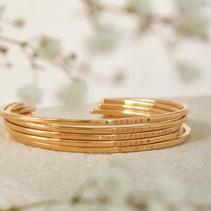 Close-up of gold stacked bangles with engraved names: Adrian, Claudia, Lucia, Leonardo, Kaylah, and Anok. Each name is separated by a small gemstone; emerald green, pink, and clear. The bangles are thin and delicate, ideal for layering.