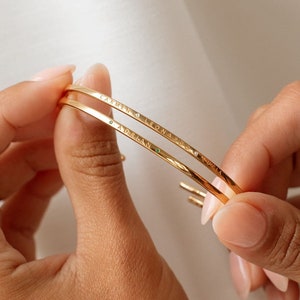 Close-up of two gold, thin, etched metal cuff bracelets. The bracelets are simple and delicate. They are shown against a white background.